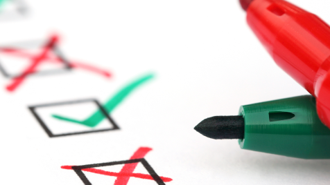 Ein grüner und ein roter Stift liegen quer übereinander auf einer Checkliste mit grünen Häkchen und roten Kreuzen